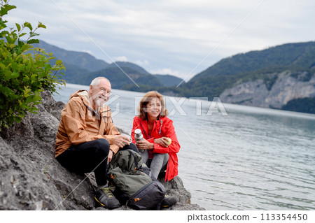 Active elderly couple hiking together in mountains. Drinking coffee and having healthy snack, to get energy. Senior tourist with backpacks resting during hike. 113354450