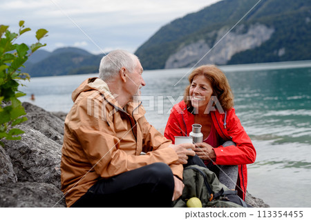Active elderly couple hiking together in mountains. Drinking coffee and having healthy snack, to get energy. Senior tourist with backpacks resting during hike. Active elderly couple hiking together in mountains. Drinking coffee and having healthy snack, to get energy. Senior tourist with backpacks resting during hike. 113354455