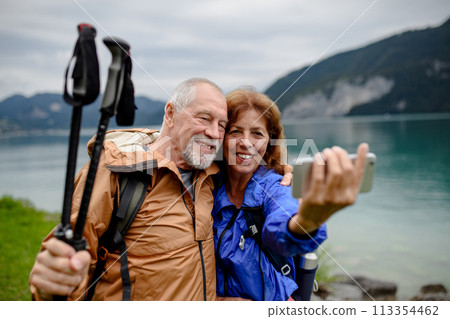 Portrait of beautiful active elderly couple taking selfie during hike in autumn mountains. Senior tourists embracing each other in front of lake Portrait of beautiful active elderly couple taking selfie during hike in autumn mountains. Senior tourists embracing each other in front of lake 113354462