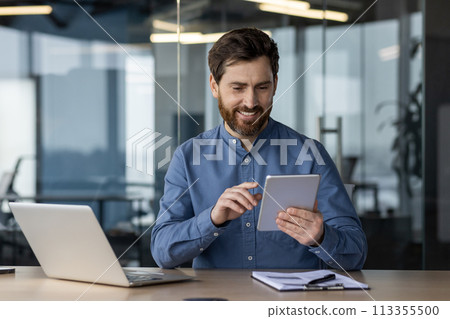 Cheerful bearded man in casual attire enjoying his tablet with a smile at an organized desk in a contemporary office environment. Cheerful bearded man in casual attire enjoying his tablet with a smile at an organized desk in a contemporary office environment. 113355500