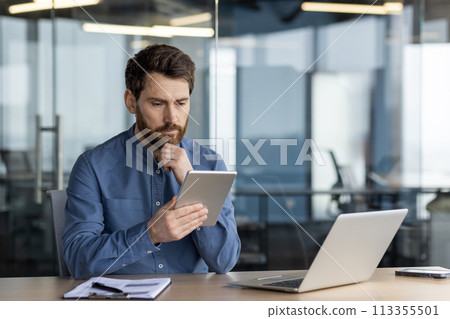 Serious male professional working on a digital tablet with a laptop and notebook by his side in a contemporary office space. Serious male professional working on a digital tablet with a laptop and notebook by his side in a contemporary office space. 113355501
