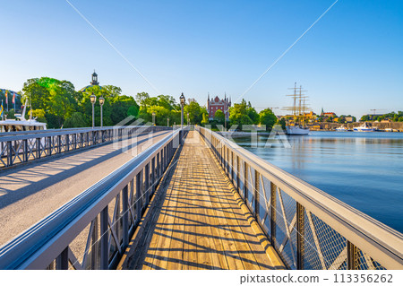 A vibrant day at Skeppsholmen Bridge in Stockholm, with clear skies and sunlight bathing the walkway, the calm water, and historic buildings in the distance. Stockholm, Sweden A vibrant day at Skeppsholmen Bridge in Stockholm, with clear skies and sunlight bathing the walkway, the calm water, and historic buildings in the distance. Stockholm, Sweden 113356262