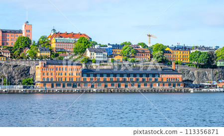 The waterfront view of Fotografiska museum in Stockholm, Sweden, basks under a clear blue sky, with vibrant buildings atop the rocky terrain in the background. 113356871