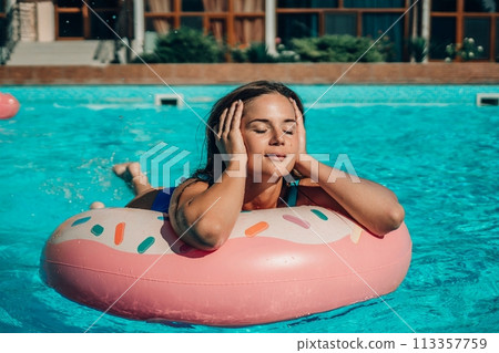 A woman is floating on a pink donut in a pool. She is wearing a blue swimsuit and she is relaxing. A woman is floating on a pink donut in a pool. She is wearing a blue swimsuit and she is relaxing. 113357759