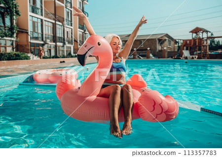 A woman is sitting on a pink inflatable flamingo in a pool. The flamingo is pink and has a blue stripe. The woman is smiling and she is enjoying herself. Concept of fun and relaxation. A woman is sitting on a pink inflatable flamingo in a pool. The flamingo is pink and has a blue stripe. The woman is smiling and she is enjoying herself. Concept of fun and relaxation. 113357783