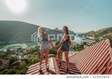 women standing on rooftop, enjoys town view and sea mountains. Peaceful rooftop relaxation. Below her, there is a town with several boats visible in the water. Rooftop vantage point. 113357819