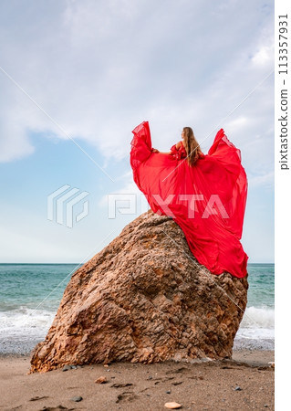 woman sea red dress. Woman with long hair on a sunny seashore in a red flowing dress, back view, silk fabric waving in the wind. Against the backdrop of the blue sky and mountains on the seashore. 113357931