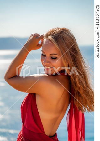 Smiling young woman in a red dress looks at the camera. A beautiful tanned girl enjoys her summer holidays at the sea. Portrait of a stylish carefree woman laughing at the ocean. 113357979