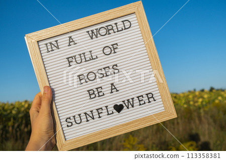 IN A WORLD FULL OF ROSES BE A SUNFLOWER text on white board next to sunflower field. Sunny summer day. Motivational caption inspirational quote. Be unique IN A WORLD FULL OF ROSES BE A SUNFLOWER text on white board next to sunflower field. Sunny summer day. Motivational caption inspirational quote. Be unique 113358381