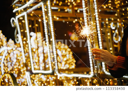 Woman holding sparkler night while celebrating Christmas outside. Dressed in a fur coat and a red headband. Blurred christmas decorations in the background. Selective focus 113358430