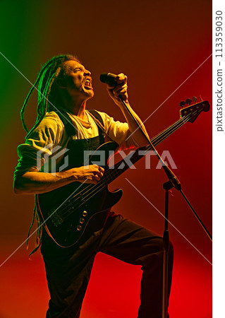 Young man, expressive singer with dreadlocks emotionally singing, performing solo against gradient red green background in neon light Young man, expressive singer with dreadlocks emotionally singing, performing solo against gradient red green background in neon light 113359030