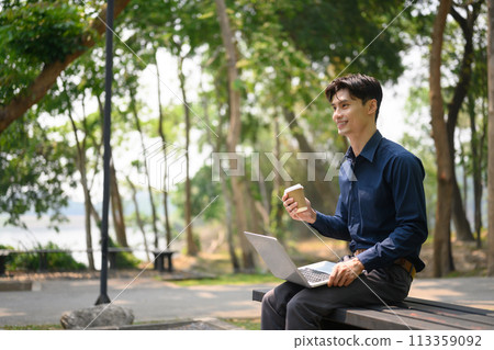 Portrait of attractive businessman sitting on a park bench and working with laptop 113359092