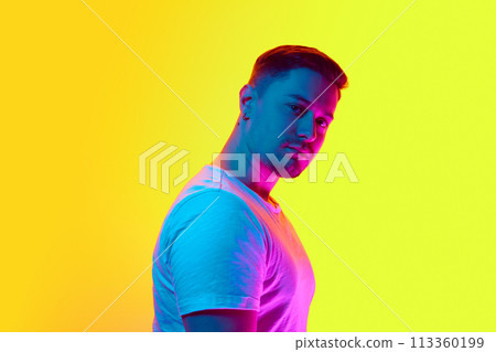 Portrait of confident young man with serious facial expression looking at camera in neon light against vibrant yellow studio background. 113360199