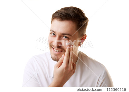 Young handsome man in T-shirt applying cream and looking with smile at camera against white studio background. Young handsome man in T-shirt applying cream and looking with smile at camera against white studio background. 113360216