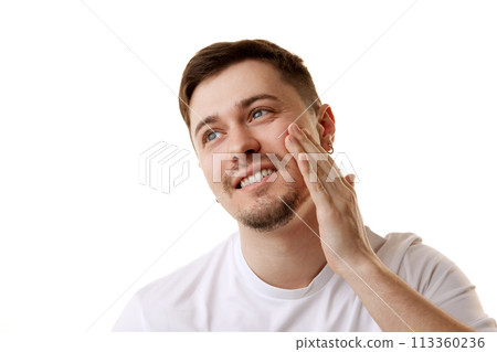 Young handsome man in white t-shirt looking away while applying facial-care serum against white studio background. Young handsome man in white t-shirt looking away while applying facial-care serum against white studio background. 113360236