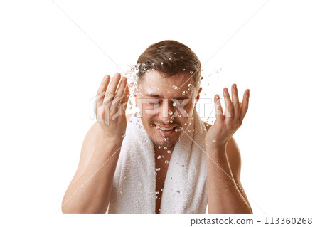 Young man with towel on shoulders washing his face with splashes of water in motion against white studio background. Young man with towel on shoulders washing his face with splashes of water in motion against white studio background. 113360268