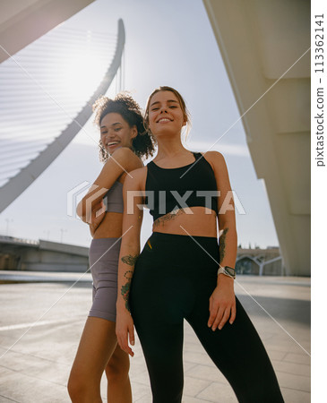 Two young female sportswomen have a rest after morning jogging standing outdoors and looks camera Two young female sportswomen have a rest after morning jogging standing outdoors and looks camera 113362141