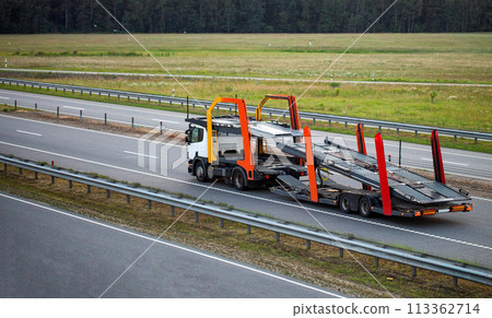 An empty tractor-trailer truck with a semi-trailer car transporter drives along the highway in the summer against the backdrop of sunset behind new cars. Concept for transporting cars from other 113362714