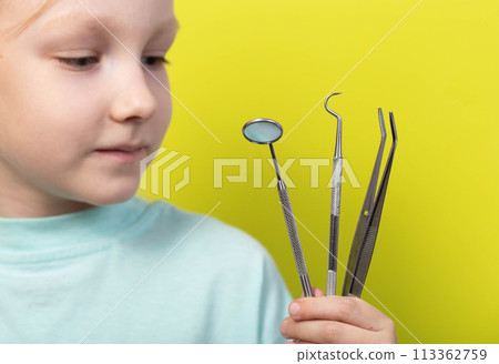 A beautiful seven-year-old girl holds dental instruments in her hand on a yellow background, close-up. The concept of therapeutic dentistry, root canal filling and teeth whitening. 113362759