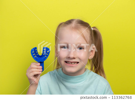 Smiling girl aged seven years old with a blue dental impression tray on a yellow background. The concept of making impressions of the dental jaw in children in orthodontics. Making veneers and 113362768