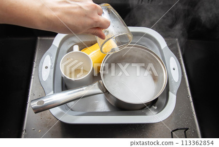 A woman's hand pours hot water into a basin with dirty dishes and environmentally friendly oxygen bleach. Washing dishes from grease and dirt. 113362854