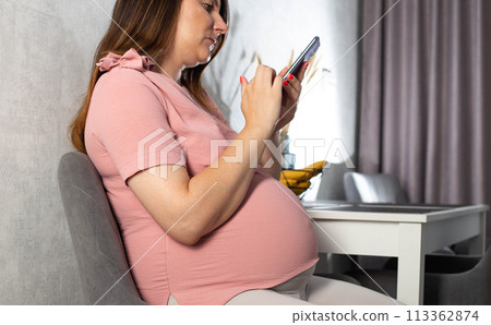 Pregnant girl with a mobile phone in the kitchen. The concept of the negative impact of phone radiation on the fetus in the womb. Pregnant girl with a mobile phone in the kitchen. The concept of the negative impact of phone radiation on the fetus in the womb. 113362874