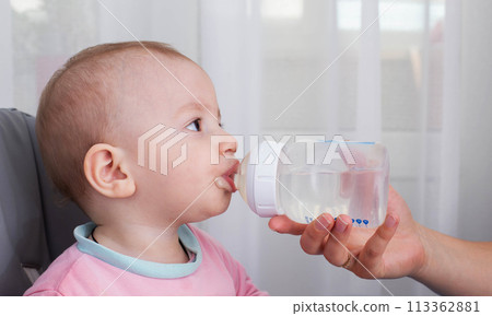 A mother's hand gives water to a baby to drink from a bottle while feeding porridge. A child boy aged one year drinks water. Copy space for text, caucasian 113362881