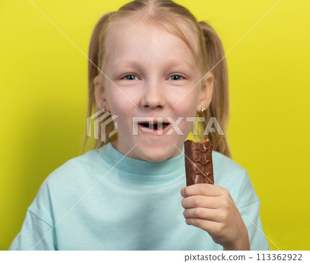 A beautiful joyful girl with white hair of seven years old eats a chocolate bar on a yellow background. Children are lovers of sweet junk food. A beautiful joyful girl with white hair of seven years old eats a chocolate bar on a yellow background. Children are lovers of sweet junk food. 113362922