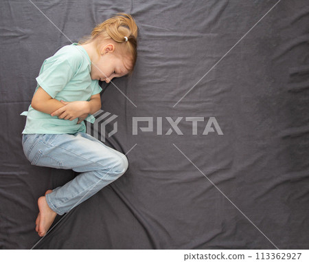 A little caucasian girl of seven years old lies on a gray bed with her legs crossed and holds her stomach with her hands. Concept of bad mood and depression in children. Onset of colds and body 113362927