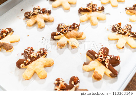 Creating snowflake-shaped cutout sugar cookies, dipped in chocolate, and adorned with crushed pecan nuts, elegantly presented on parchment. 113363149