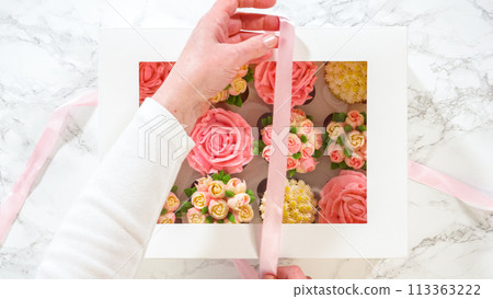 Flat lay. In a woman's hands, a white paper box filled with gourmet cupcakes, each adorned with vibrant roses and tulips crafted from buttercream frosting, is gently being closed. 113363222