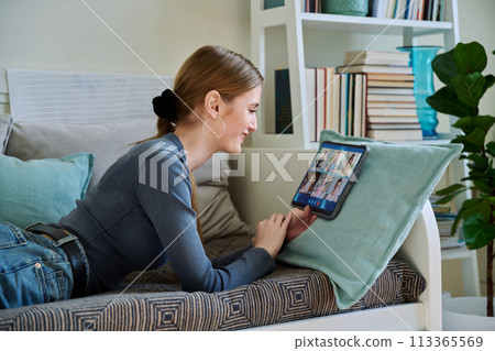 Female teenage student having group video conference, using digital tablet, lying on couch at home. Female teenage student having group video conference, using digital tablet, lying on couch at home. 113365569