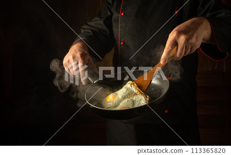 A chef in uniform holds a frying pan with a hot fried egg and a wooden spatula in his hands. Low key concept of cooking scrambled eggs. Place for advertising on a black background 113365820