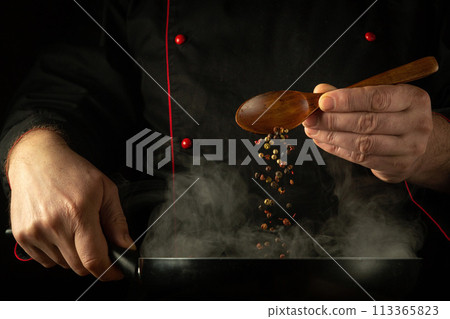 The cook adds dry pepper with a spoon into a hot frying pan with steam. Low key concept of preparing a delicious dinner in the saloon kitchen. European cuisine 113365823