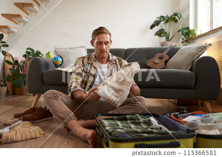 Travelling and tourism concept. Happy young man, sits on floor with luggage opened, packing suitcase for holiday, going on vacation, putting clothes inside a bag Travelling and tourism concept. Happy young man, sits on floor with luggage opened, packing suitcase for holiday, going on vacation, putting clothes inside a bag 113366581