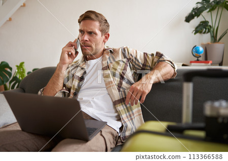 Portrait of young man with credit card, sitting and using laptop, making a phone call, booking hotel or tickets, confirming his purchase over telephone, has suitcase near him 113366588