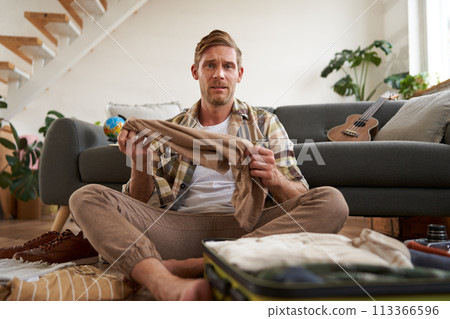 Portrait of confused man, sitting in living room with suitcase, holding pair of trousers, looking clueless, struggling to pack for holiday Portrait of confused man, sitting in living room with suitcase, holding pair of trousers, looking clueless, struggling to pack for holiday 113366596