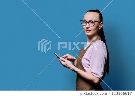 Portrait of 30s woman in apron with smartphone looking at camera on blue background 113366617