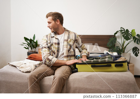 Tourism and travelling concept. Portrait of handsome young man going on holiday, packing his suitcase, checking luggage, sitting on bed and looking at packed things Tourism and travelling concept. Portrait of handsome young man going on holiday, packing his suitcase, checking luggage, sitting on bed and looking at packed things 113366887