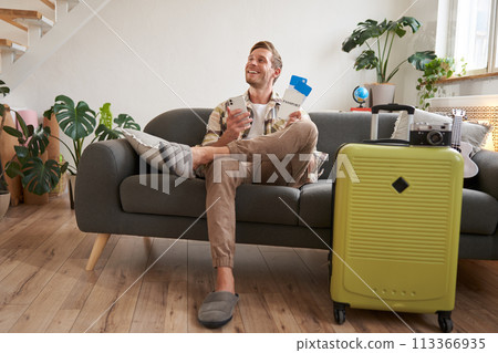 Handsome young man sits on sofa with suitcase, holds smartphone, books hostel or holiday vacation, chats on mobile phone 113366935
