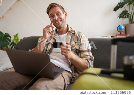 Portrait of young man with credit card, sitting and using laptop, making a phone call, booking hotel or tickets, confirming his purchase over telephone, has suitcase near him 113366956