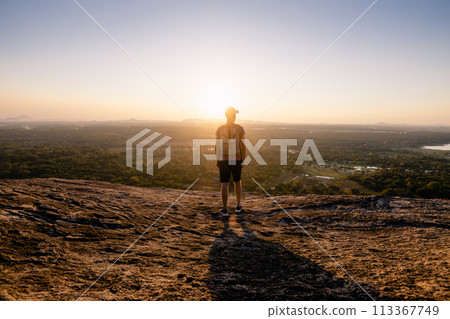 Traveler on rock overlooking sunset over tropical landscape. Traveler on rock overlooking sunset over tropical landscape. 113367749