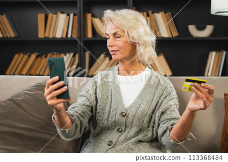 Happy middle aged woman shopping online with smartphone paying with credit card. Mature lady buying on Internet enter credit card details at home indoors. Online shopping delivery service ecommerce 113368484