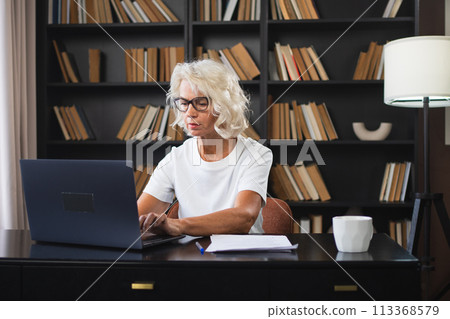 Confident stylish middle aged senior woman using laptop at workplace. Older mature lady businesswoman sitting at office table with laptop and paper document. Boss leader teacher professional worker Confident stylish middle aged senior woman using laptop at workplace. Older mature lady businesswoman sitting at office table with laptop and paper document. Boss leader teacher professional worker 113368579