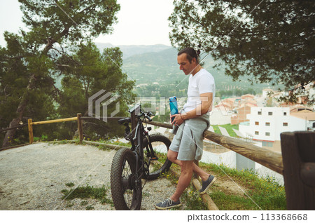 Cyclist, sportsman holding a bottle of water, having a break while riding using a motor-powered electric bicycle, a bike with pedal-assist system. Active people, nature and healthy lifestyle concept Cyclist, sportsman holding a bottle of water, having a break while riding using a motor-powered electric bicycle, a bike with pedal-assist system. Active people, nature and healthy lifestyle concept 113368668