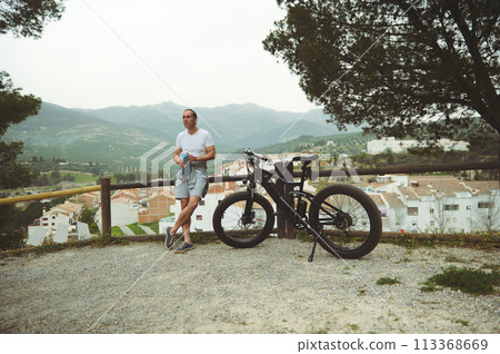 Full length portrait of a young man cyclist, standing nearby his electric bike, relaxing after biking in mountains Full length portrait of a young man cyclist, standing nearby his electric bike, relaxing after biking in mountains 113368669