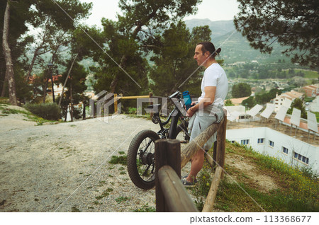 Rear view of handsome active young man, a cyclist in electric bike, relaxing after biking in mountains. Using e-bike as eco-friendly mode of transport for trip or adventure. Electronic mobility Rear view of handsome active young man, a cyclist in electric bike, relaxing after biking in mountains. Using e-bike as eco-friendly mode of transport for trip or adventure. Electronic mobility 113368677
