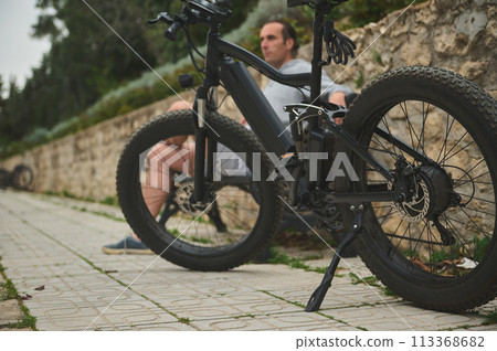 Selective focus on an innovated modern black electric bike, parked near a bench with a sitting cyclist 113368682