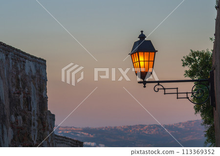 A beautiful street lamp lights up against the background of a remote hilly residential development at dusk. Symbol of the onset of night or morning 113369352