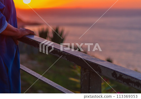 Handrail on which an elderly unrecognizable man leans with his hands stands against the background of an amazing sunset over the sea horizon, enjoying the magnificent landscape, view and life Handrail on which an elderly unrecognizable man leans with his hands stands against the background of an amazing sunset over the sea horizon, enjoying the magnificent landscape, view and life 113369353